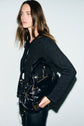 A woman with long brown hair wears a gilet noir—a black, textured jacket adorned with subtle lurex sequins—and holds a shiny black handbag with gold hardware, standing against a plain light background.