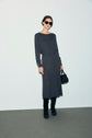 Woman wearing sunglasses, a dark ribbed long-sleeve dress with boutons dorés and subtle maille fantaisie details, black knee-high boots, and holding a small black handbag stands against a plain background.
