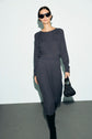 A woman in dark sunglasses wears a ribbed, long-sleeve, charcoal knit dress with boutons dorés and black boots, holding a black textured handbag, walking against a light background.