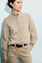 A person wears a beige pull à col montant with boutons argentés, tucked into light tan pants with a braided dark belt, standing against a pale blue background.