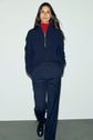 A person stands against a plain background wearing a navy pull col camionneur in maille texturée, a red turtleneck underneath, navy pants, and black shoes, with hands in pockets and a relaxed expression.