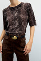 Person wearing a brown snake-print short-sleeve top featuring un délicat imprimé python, brown pants with front pockets, and a black belt with a large gold oval buckle, standing against a light background.
