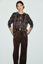 A person stands against a light background wearing a brown imprimé python sweater, reminiscent of motif animalier, paired with dark brown suede pants, hands in pockets and a neutral expression.