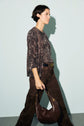 Woman with short dark hair walks side-on against a plain background, wearing an imprimé python top, brown pants, and carrying a dark brown handbag in her left hand.