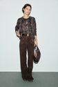 Woman standing against a light background, wearing a brown blouse with motif animalier snakeskin patterns, dark brown wide-leg pants, and holding a large brown handbag. She has short dark hair and a neutral expression.
