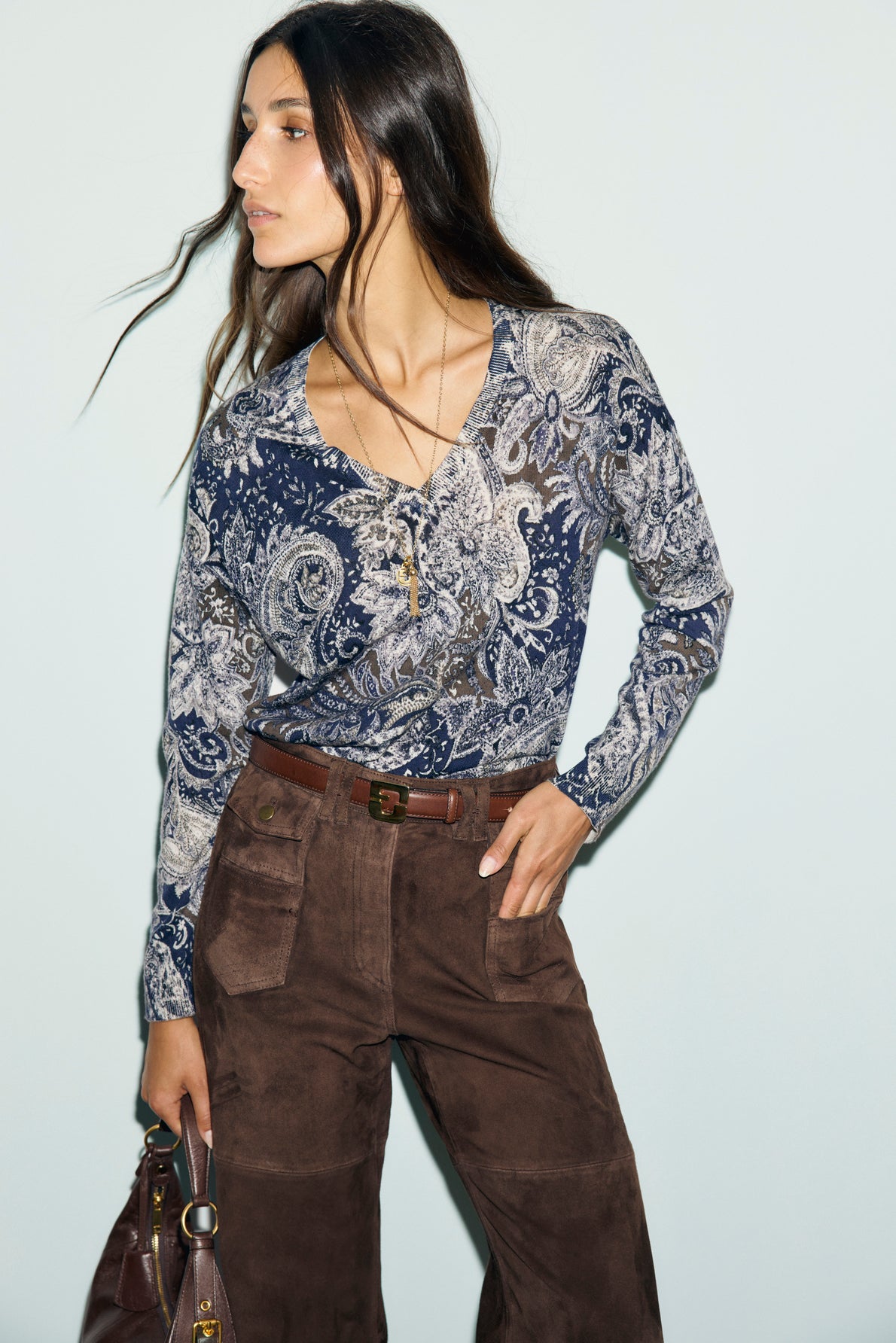 Woman with long wavy hair wearing a blue and white imprimé cachemire top and brown high-waisted pants with front pockets, holding a brown handbag, looking to the side against a light background.