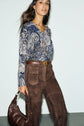 Woman wearing a blue imprimé cachemire top and brown suede pants, holding a brown handbag, walking confidently with her hair flowing and a slight smile.