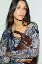 Woman with long brown hair wearing a blue imprimé cachemire top holds a brown leather purse against her chest, looking at the camera with a neutral expression.