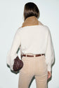 A woman stands facing away, wearing a pull écru sweater with a tan collar, beige pants, and a brown belt. She holds a small textured brown handbag in her left hand.