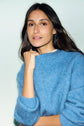A woman with long dark hair wears a soft blue sweater, gazing slightly to the side with a relaxed expression. Her hand is gently holding the collar of her pull laine mohair. The background is a light, neutral color.
