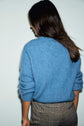 A person with long dark hair wearing a pull bleu intense and houndstooth-patterned pants stands facing away from the camera against a plain light background.