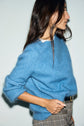Woman with long brown hair wearing a pull bleu intense in fuzzy mohair and plaid pants, standing sideways with one hand in her pocket against a plain light background.