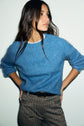 A person with long brown hair wears a pull bleu intense—a fluffy, poilu sweater made of laine mohair—over a white shirt and checkered pants, standing with hands on hips and looking to the side against a plain light background.