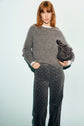 A woman with shoulder-length auburn hair wears a gray pull col rond mohair sweater over a white shirt, patterned dark pants, and holds a black handbag, standing against a light background.