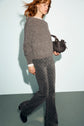 A woman with red hair, wearing a gray pull col rond fuzzy sweater and patterned gray pants, carries a small black handbag and walks against a light background.