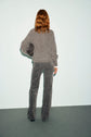 A person with wavy, shoulder-length hair stands facing a light blue wall, wearing a pull épais laine gray sweater and patterned gray pants, holding a black handbag under their left arm.