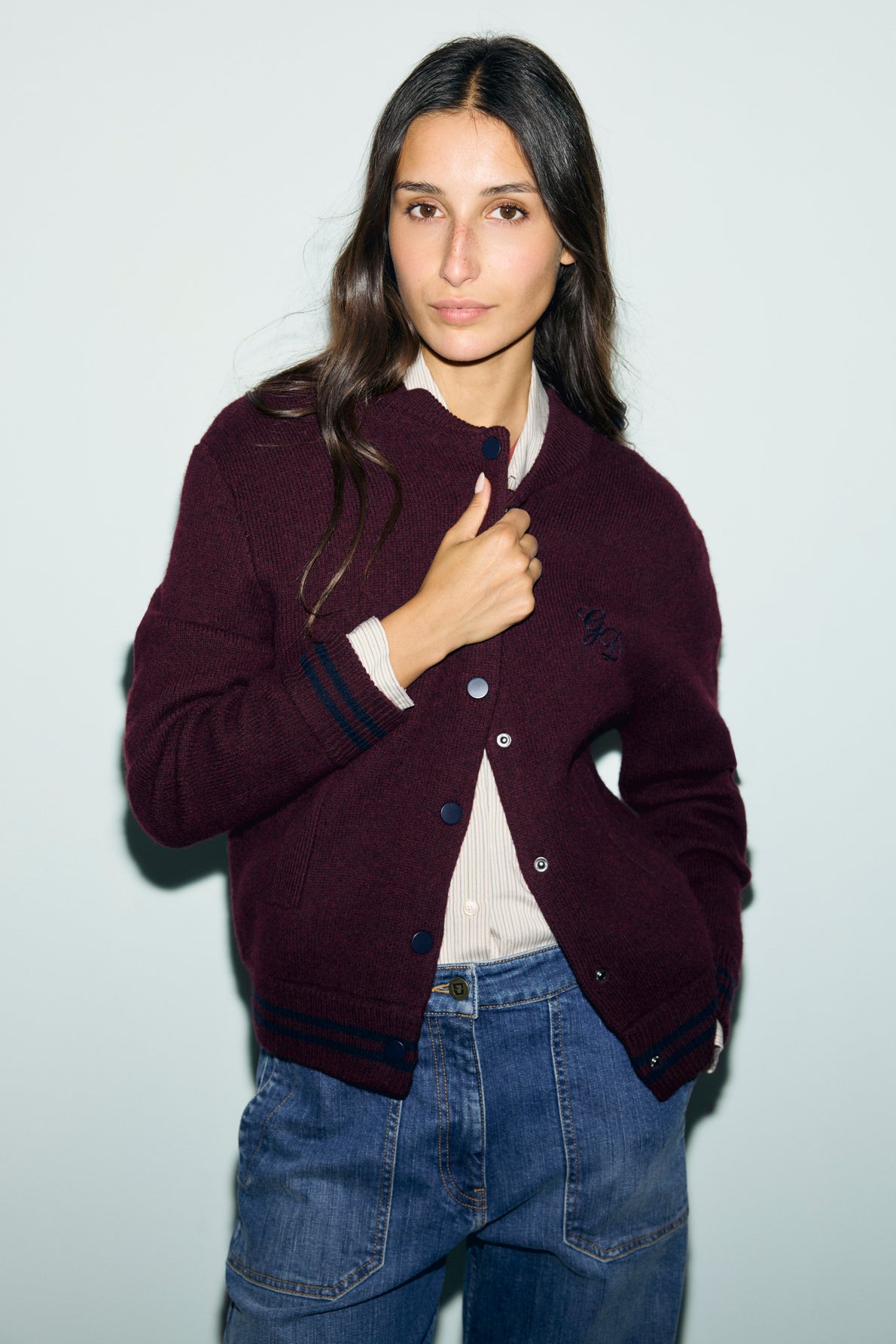 A woman with long dark hair wears a bordeaux cardigan over a light blouse and blue jeans, standing against a plain light background and looking at the camera with a slight smile.