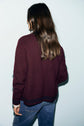 A person with long brown hair, wearing a maroon gilet en laine and blue jeans, stands facing away from the camera against a plain light background.