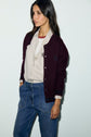A woman with long dark hair wears a bordeaux cardigan over a striped button-up shirt and high-waisted blue jeans, standing against a plain light background.