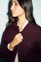 A person in a bordeaux cardigan with navy embroidery over a striped shirt looks to the side, holding the sweater closed with one hand against a plain background.