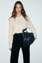Woman with long brown hair wears a pull écru in soft laine alpaga and high-waisted black pants, holding a large black fringed bag against a plain white background.