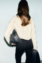 A woman with long brown hair, wearing a pull écru in laine alpaga featuring a subtle motif losanges and black pants, stands facing away. She holds a black fringed handbag and a black jacket.
