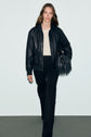 A woman with long brown hair wears a black leather jacket, a pull écru en laine alpaga à motif losanges, black pants, and holds a black fringed handbag while walking toward the camera against a plain background.