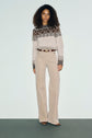 Woman with curly hair wears a beige and brown patterned sweater in motif Fair Isle, beige high-waisted corduroy pants, and brown shoes. She stands against a plain light-colored background, looking directly at the camera.