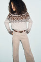 Person with curly hair wearing a beige and brown patterned sweater en laine alpaga with a motif Fair Isle and light-colored corduroy pants, looking to the side against a pale blue background.
