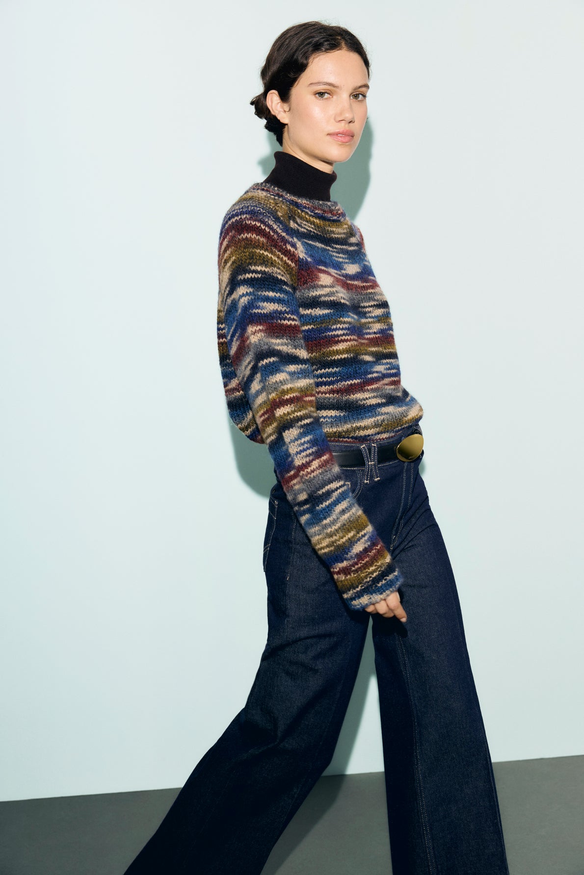 A person wearing a multicolored pull rayé en laine mélangée, dark wide-leg jeans, and a black turtleneck stands against a pale background, looking toward the camera with a neutral expression.
