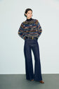 A person stands against a plain background wearing a colorful pull rayé sweater in laine mélangée, high-waisted dark bell-bottom jeans, a wide belt, and brown shoes, with hands behind their back.
