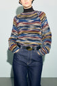 Person wearing a multicolored, pull rayé en laine mélangée over a dark turtleneck and high-waisted blue jeans with a black belt, hands in pockets, standing against a plain light background.