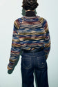 A person with short dark hair, seen from behind, wears a colorful pull rayé and dark blue jeans with white stitching, standing against a light blue background.