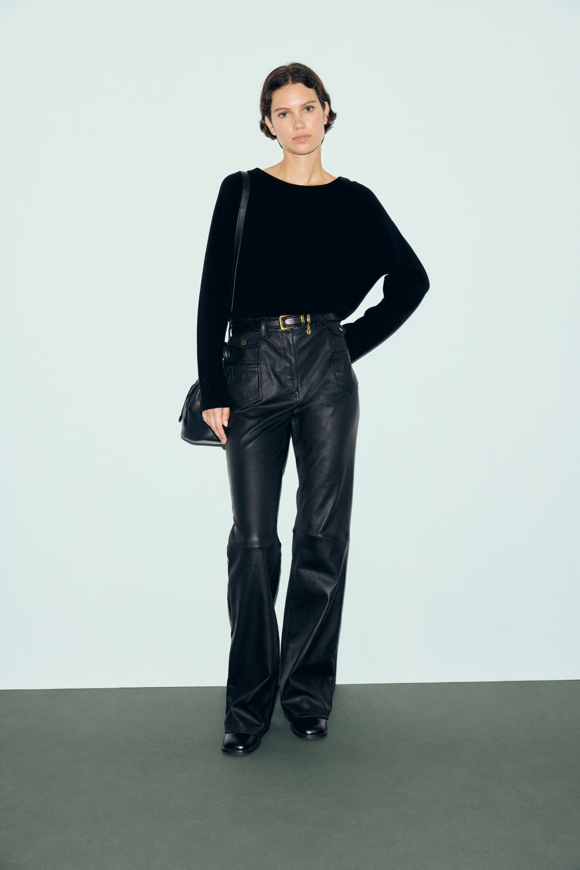 A person stands against a plain background, wearing a chic pull noir with delicate strass embellishments, paired with high-waisted black leather pants cinched by a belt, black shoes, and carrying a black shoulder bag.