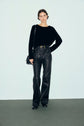 A person stands against a plain background, wearing a chic pull noir with delicate strass embellishments, paired with high-waisted black leather pants cinched by a belt, black shoes, and carrying a black shoulder bag.