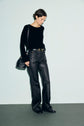 A person wearing a pull noir in soft laine, black leather pants, and black shoes stands against a light background, looking down. They carry a black shoulder bag adorned with subtle strass details and have short dark hair.