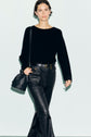 A person wearing a pull noir en laine, black leather pants with pockets, and carrying a black shoulder bag embellished with strass stands against a pale background.