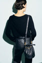 A person with short dark hair, dressed in a pull noir en laine adorned with subtle strass and black leather pants, stands facing away. They carry a black leather bucket bag with gold accents over their shoulder against a plain light blue background.
