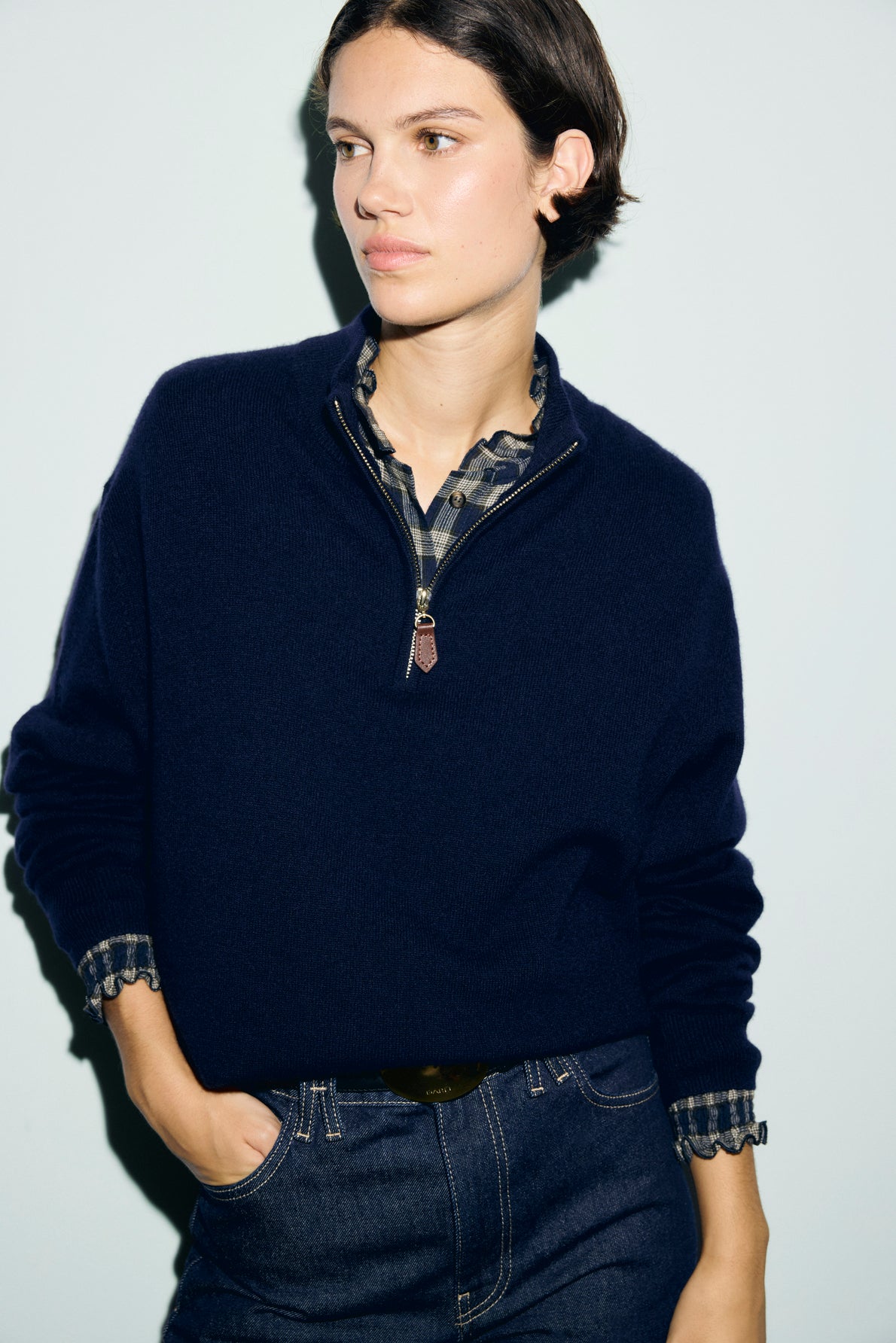 A person in a cachemire bleu marine sweater over a plaid shirt stands against a light background with one hand in their jean pocket, looking to the side.