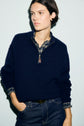 A person in a cachemire bleu marine sweater over a plaid shirt stands against a light background with one hand in their jean pocket, looking to the side.