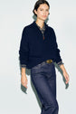 A person stands against a light background, wearing a pull camionneur en cachemire bleu marine à col montant over a plaid shirt and dark blue jeans. They have short, dark hair and are looking confidently at the camera.