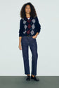 A person with curly hair wears a polo bleu marine à motif losanges over a maroon top, blue jeans, and black loafers, standing against a plain light background.