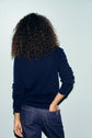 Person with curly hair, wearing a polo bleu marine en laine and blue jeans, stands facing away from the camera with one hand in their back pocket against a light blue background. Subtle motif losanges add texture to the sweater’s deep navy hue.