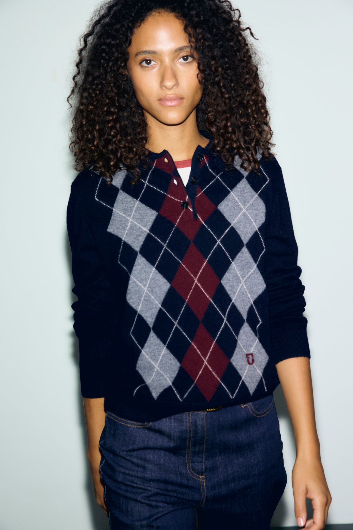 A person with curly hair wears a polo bleu marine en laine featuring a red and gray motif losanges, layered over a white shirt and paired with dark jeans, standing against a light background.