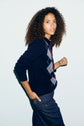 A person with curly hair wears a polo laine bleu marine à motif losanges (navy wool polo sweater with a diamond pattern) over a plaid shirt and dark jeans, posing with one hand in their pocket and looking confidently at the camera against a plain light background.