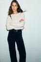 Woman with long brown hair wears a pull torsades en cachemire—a white knit sweater with an esprit rustique chic—paired with dark velvet pants and a belt, standing against a plain light background with arms crossed and a slight smile.