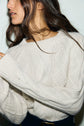 A woman with long brown hair wears a chunky, cream-colored pull torsades, embodying un esprit rustique chic. Only her lower face and upper body are visible as she stands against a plain light background.