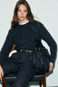 Woman with long brown hair sits on a chair, wearing a dark knit sweater with delicate torsades and pants, with a black belt and holding a black woven handbag, looking directly at the camera.