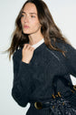A woman with long brown hair wears a dark knit sweater featuring délicate torsades, a black belt with a gold buckle, and holds a black handbag, standing against a plain light background that complements her esprit irlandais style.