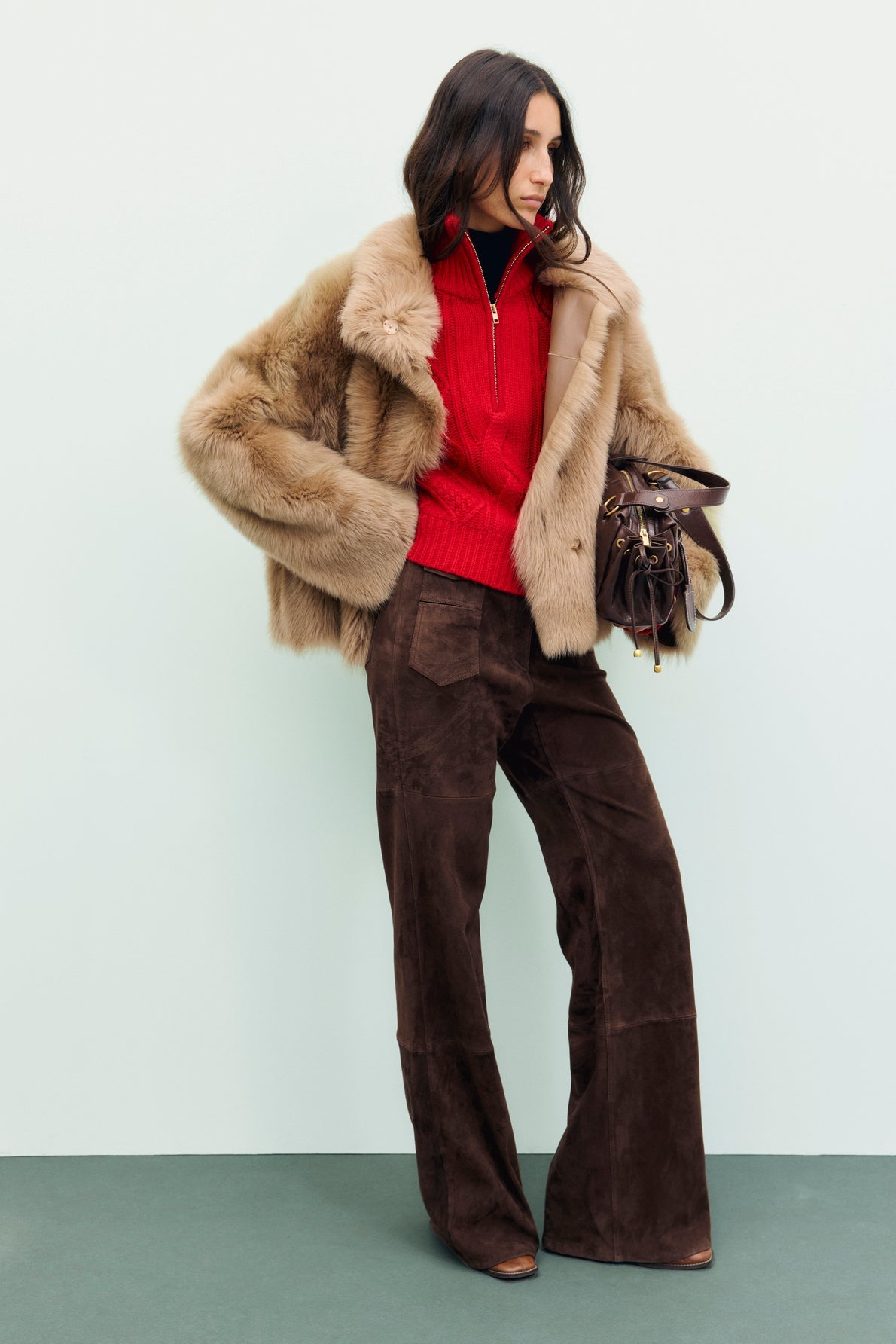 Woman in a tan fur jacket, red pull camionneur laine with col montant zippé, and brown wide-leg pants stands holding a brown handbag against a pale background. Her hair is loose, and she looks to the side.